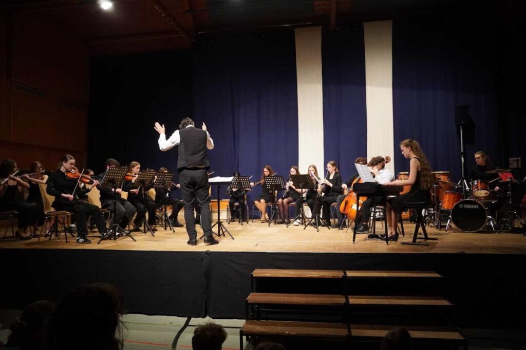 Orchesteraufführung mit Dirigent auf der Bühne rechts.